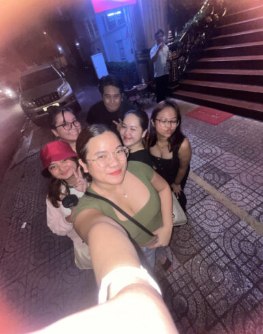 Group selfie of six friends smiling on a city sidewalk at night, with bright lights and stairs in the background.