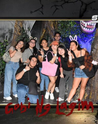 Group of friends posing in a Halloween-themed photo booth at Star City’s 'Gabi Ng Lagim' attraction, surrounded by spooky decorations like trees, bats, and a full moon backdrop.