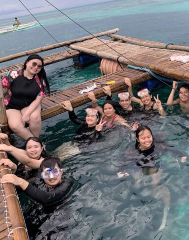 Group of people standing in shallow water beneath a large rock formation with a natural arch, enjoying a scenic beach location.