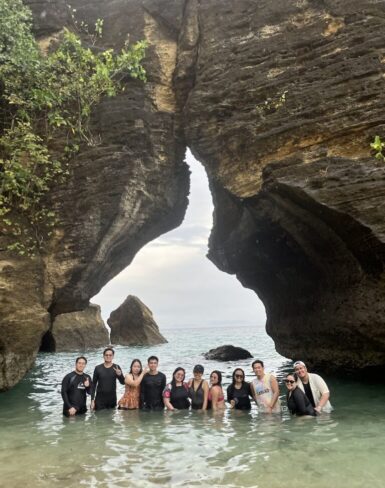 Group of people standing in shallow water beneath a large rock formation with a natural arch, enjoying a scenic beach location.