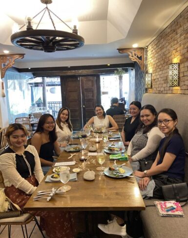 Group of women seated around a dining table at a restaurant, smiling and enjoying a meal together in a warmly lit setting with rustic decor.